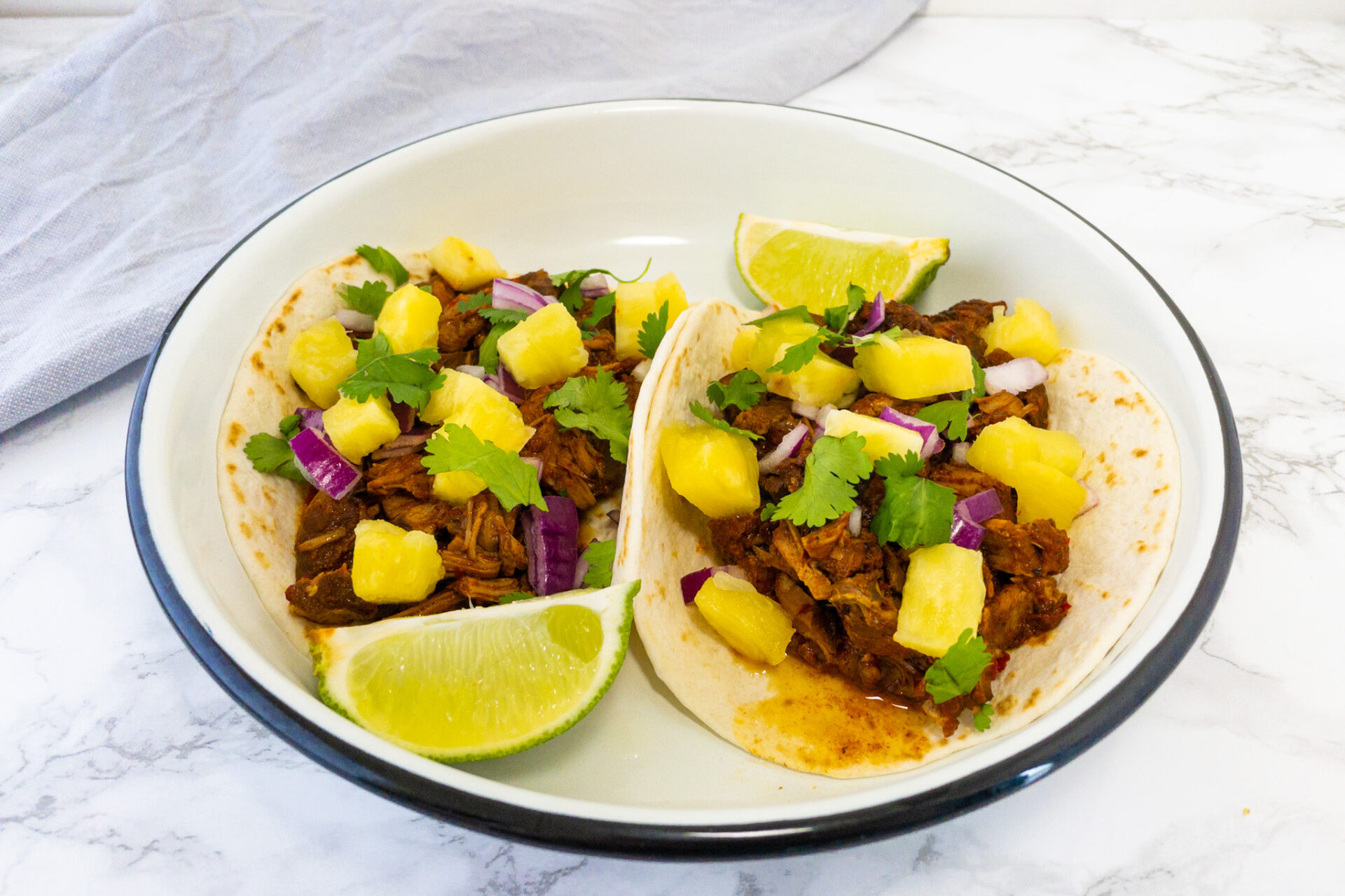 Taco's al pastor op een wit bord met daarbij limoenen en blokjes ananas