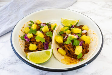 Taco's al pastor op een wit bord met daarbij limoenen en blokjes ananas